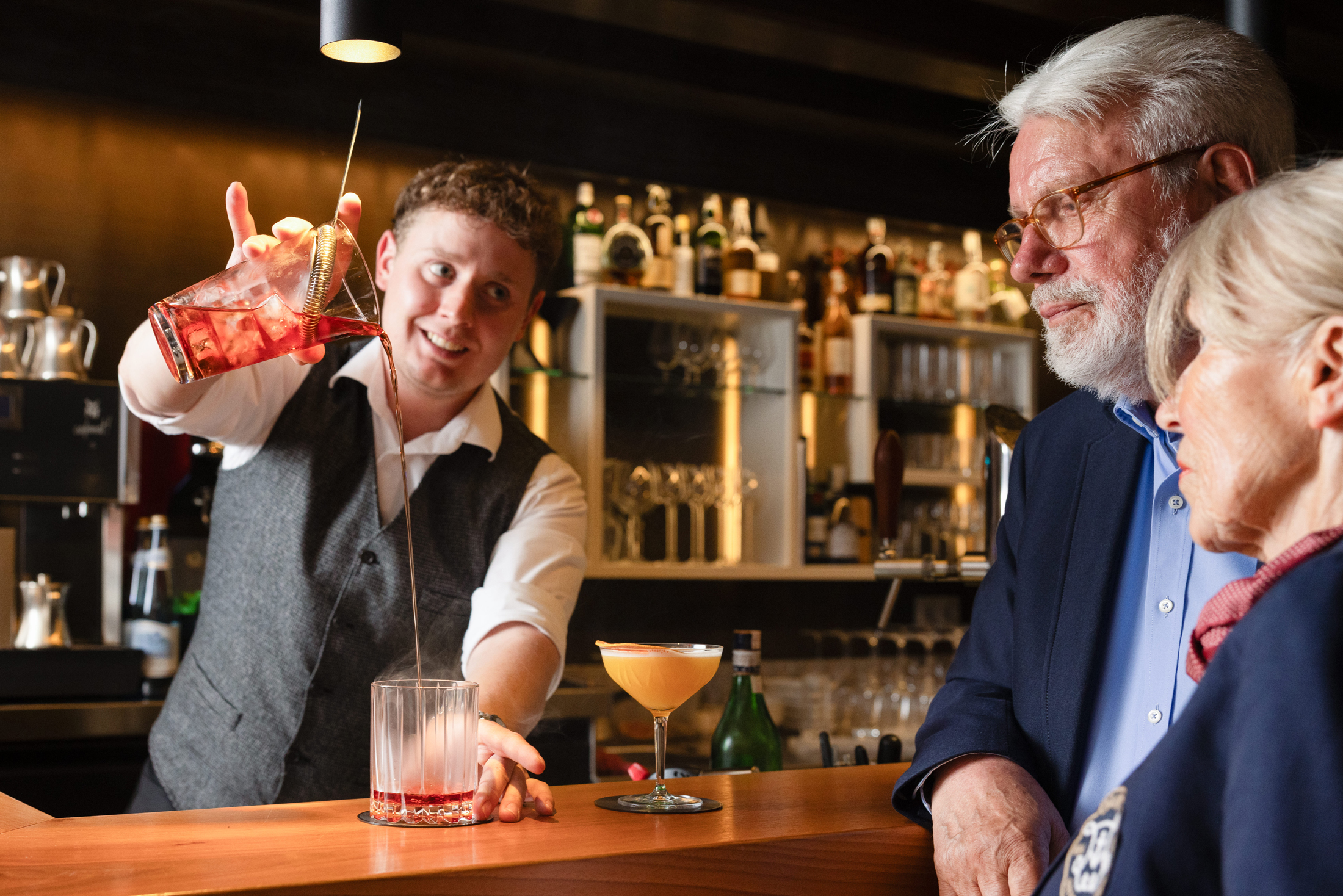 Barkeeper gießt Cocktail über Eis; zwei bunte Drinks auf Tresen; älterer Mann im Sakko beobachtet, Flaschenregal hinten.