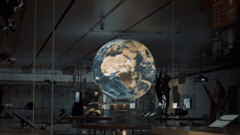 Gro&szlig;e, leuchtende Erdkugel als Ausstellungsinstallation in dunklem Innenraum; Personen, Ger&uuml;ste und Technik im Raum.