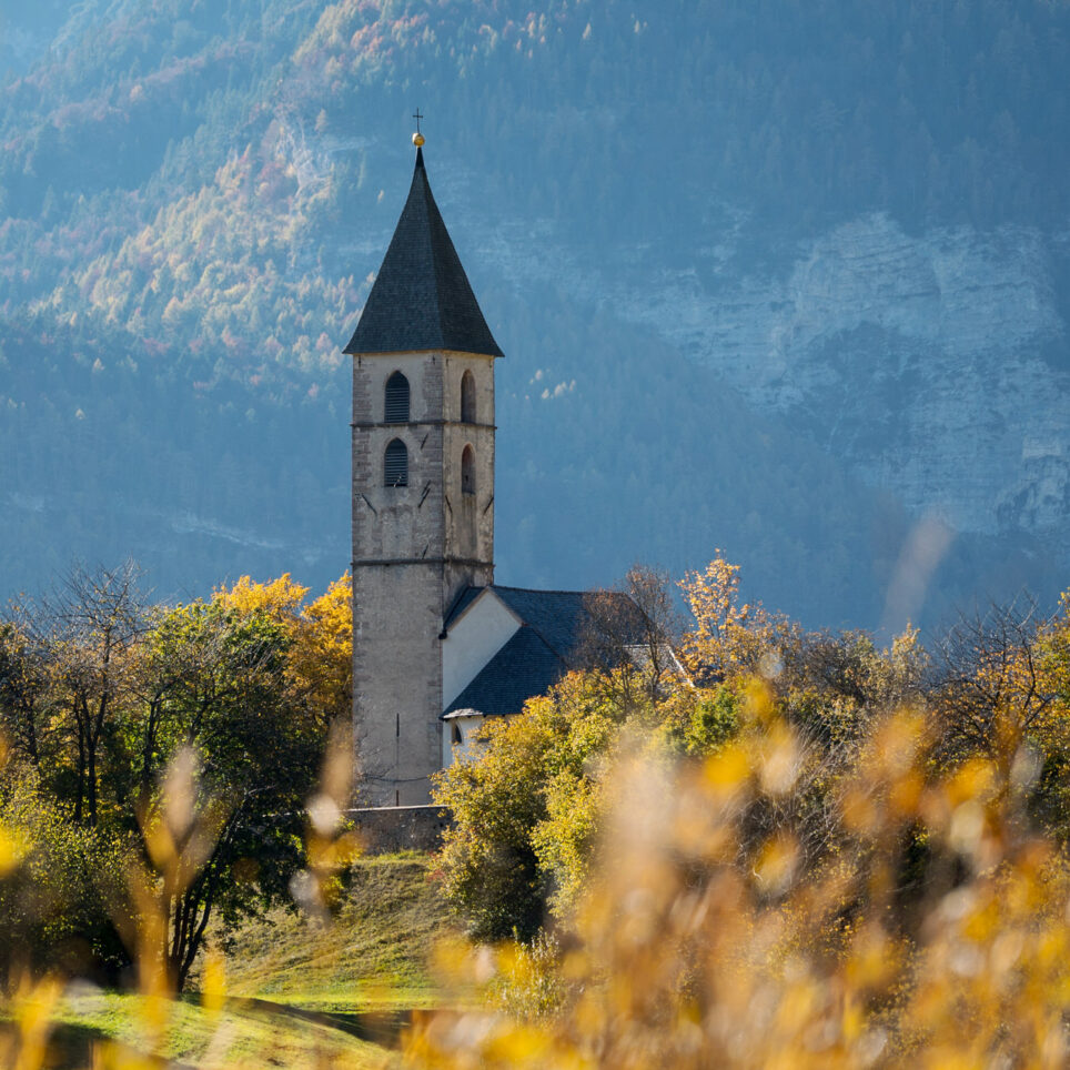 Kirchturm in Weinlandschaft Südtirols, dahinter bewaldete Hänge und felsige Berggipfel