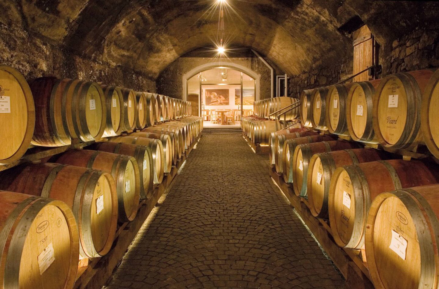 Gewölbter Weinkeller mit symmetrischen Reihen von Holzfässern entlang eines beleuchteten Mittelgangs und steiniger Decke.