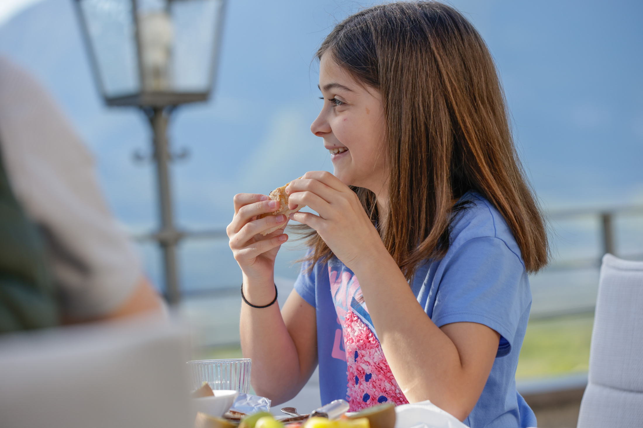 Person sitzt lächelnd an einem Außentisch im Restaurant am Kalterer See, hält ein Sandwich; Teller und Glas vorn.