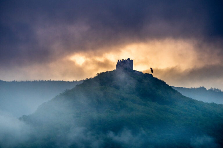 Burgruine auf einem nebelverhangenen Hügel, dramatische Wolken und Lichtschein am Horizont