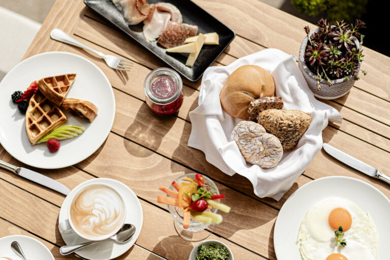 Frühstück auf Holztisch: Croissants, Körnerbrötchen in Serviette, Marmelade, Cappuccino, kleines Dessertglas und Besteck.