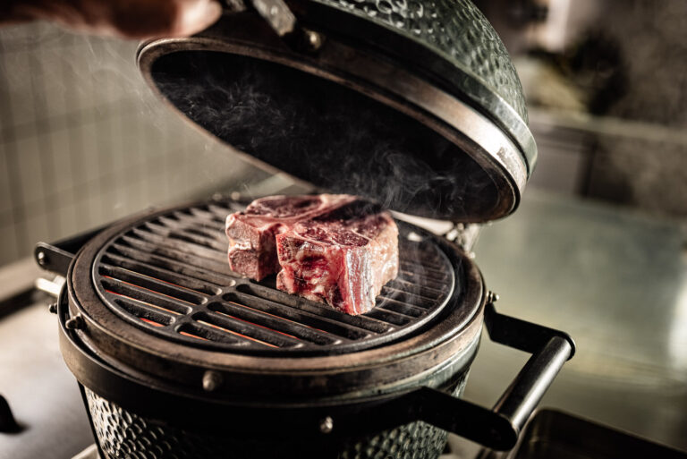 Zwei dicke rohe Steaks auf dem Rost eines geöffneten Keramikgrills, Glut und geöffneter Deckel im Hintergrund