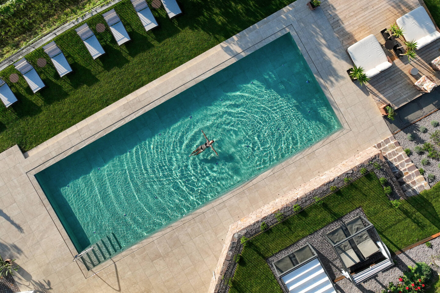 Rechteckiger Außenpool eines Hotels, eine Person treibt auf dem Rücken im Wasser; Liegestühle und Rasen an einer Seite.