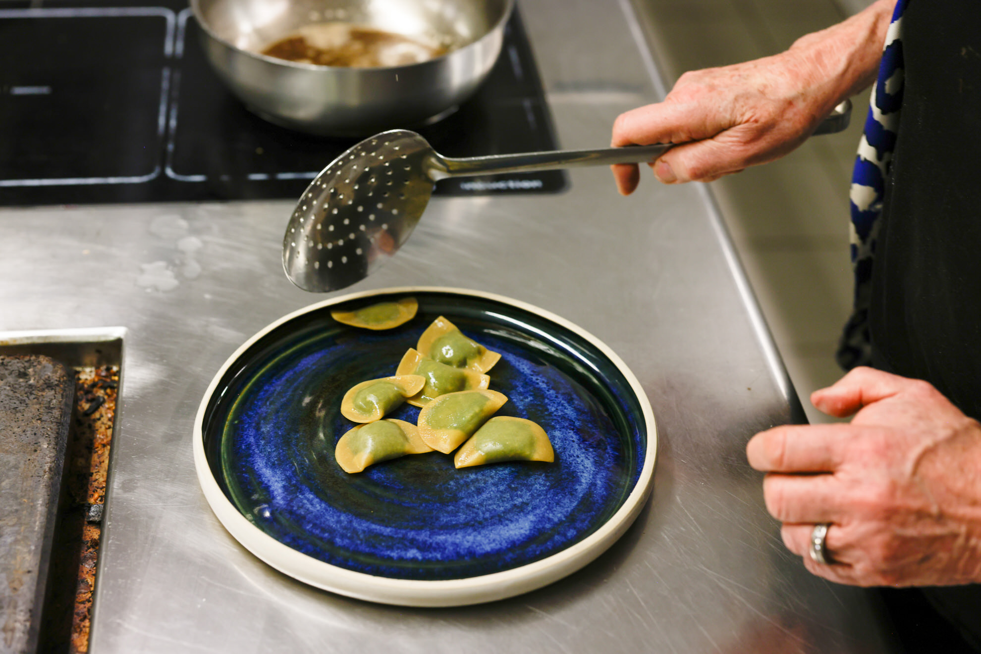 Eine Hand setzt mit Schauml&ouml;ffel drei gr&uuml;nliche Schlutzkrapfen auf blauen Teller; Edelstahlkochfeld und Pfanne im Hintergrund
