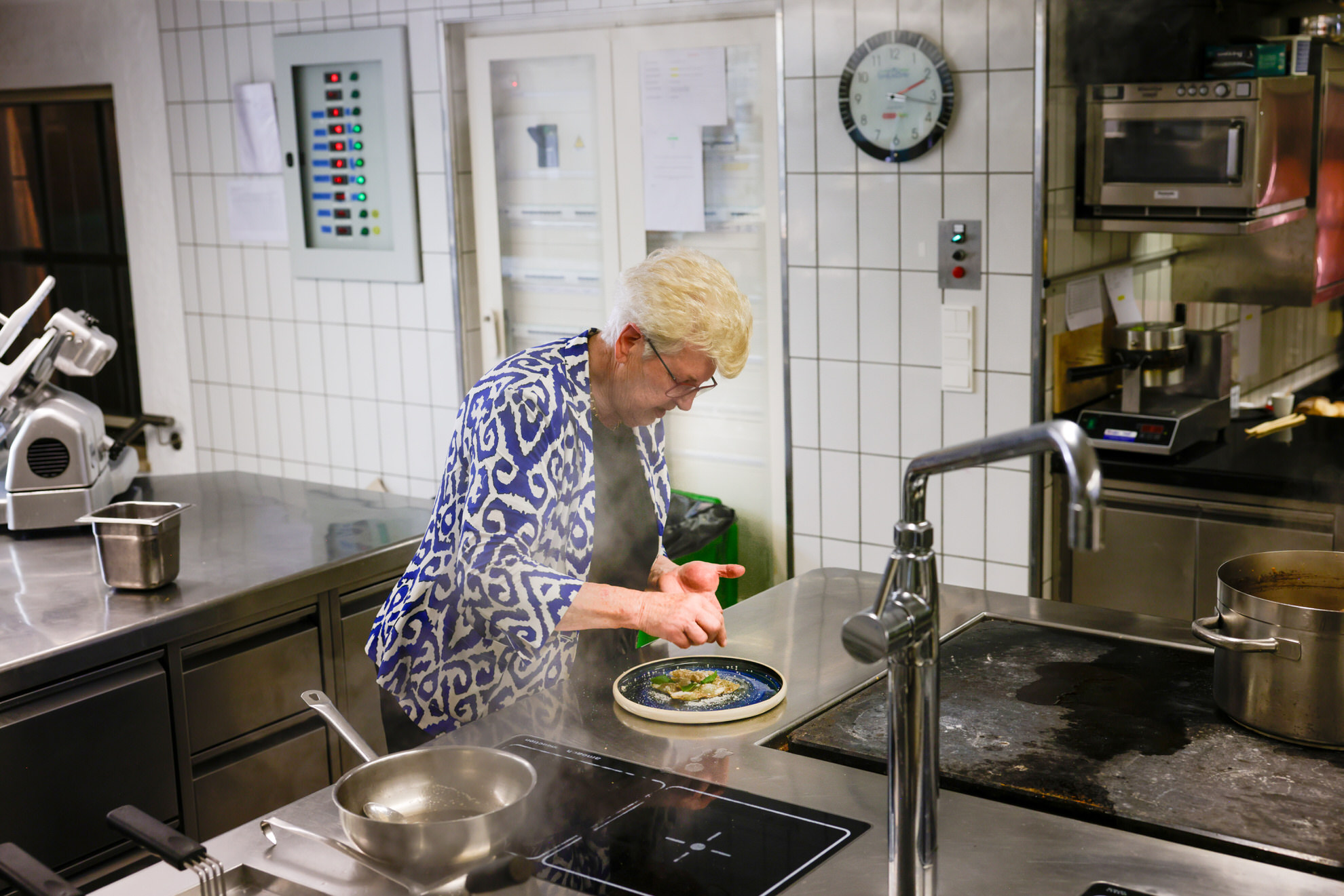 Person in gemusterter Jacke und Kopftuch r&uuml;hrt in Pfanne am Herd in gefliester K&uuml;che; Wanduhr, Sp&uuml;le und T&ouml;pfe sichtbar.