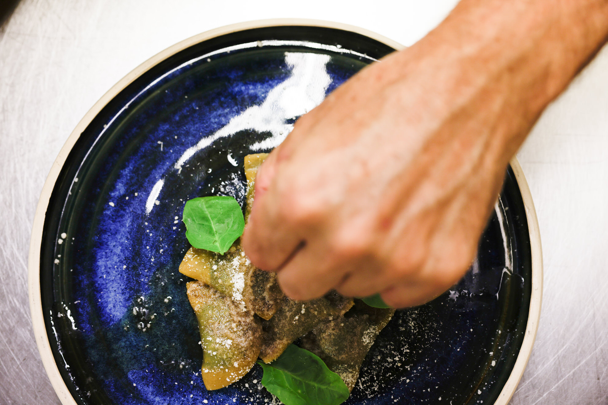 Hand streut Gew&uuml;rz &uuml;ber drei Schlutzkrapfen auf blauem Teller, dazu ein gr&uuml;nes Kr&auml;uterblatt