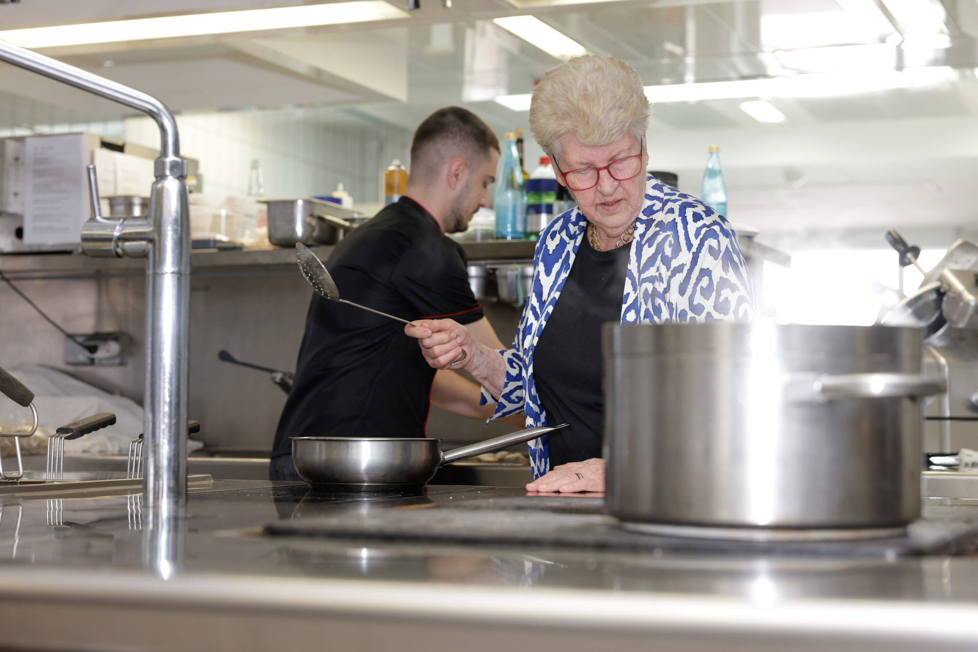 &Auml;ltere Frau und Koch in Gro&szlig;k&uuml;che am Herd; Frau r&uuml;hrt in Pfanne, gro&szlig;er Kochtopf im Vordergrund.