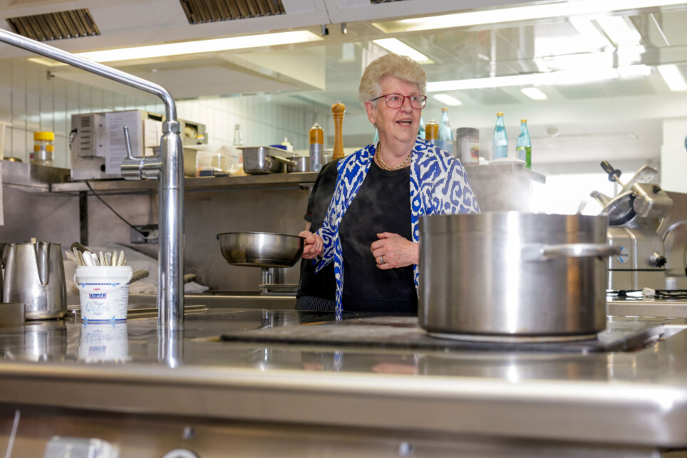 Ältere Frau mit kurzer blonder Frisur und Brille in Küchenkleidung steht in Hotelküche vor großen Edelstahltöpfen.