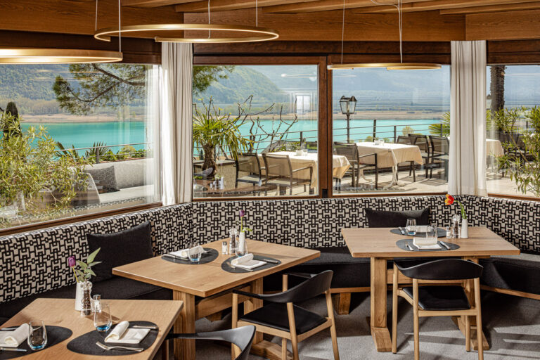 Hotelrestaurant mit Holztischen, gepolsterten Sitzbänken, großen Panoramafenstern und Terrasse mit Blick auf Kalterer See.