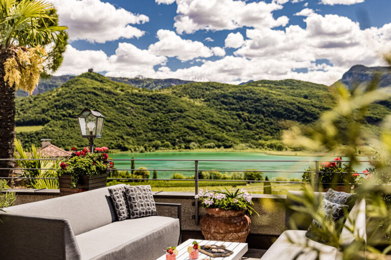Terrassenbereich mit Sitzbank, Pflanzkästen und Blick auf den Kalterer See, grüner Hang und wolkiger Himmel.