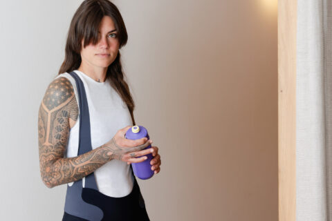 Frau mit schulterlangem Haar und Armt&auml;towierungen h&auml;lt eine lila Trinkflasche vor heller Wand und Wandlampe