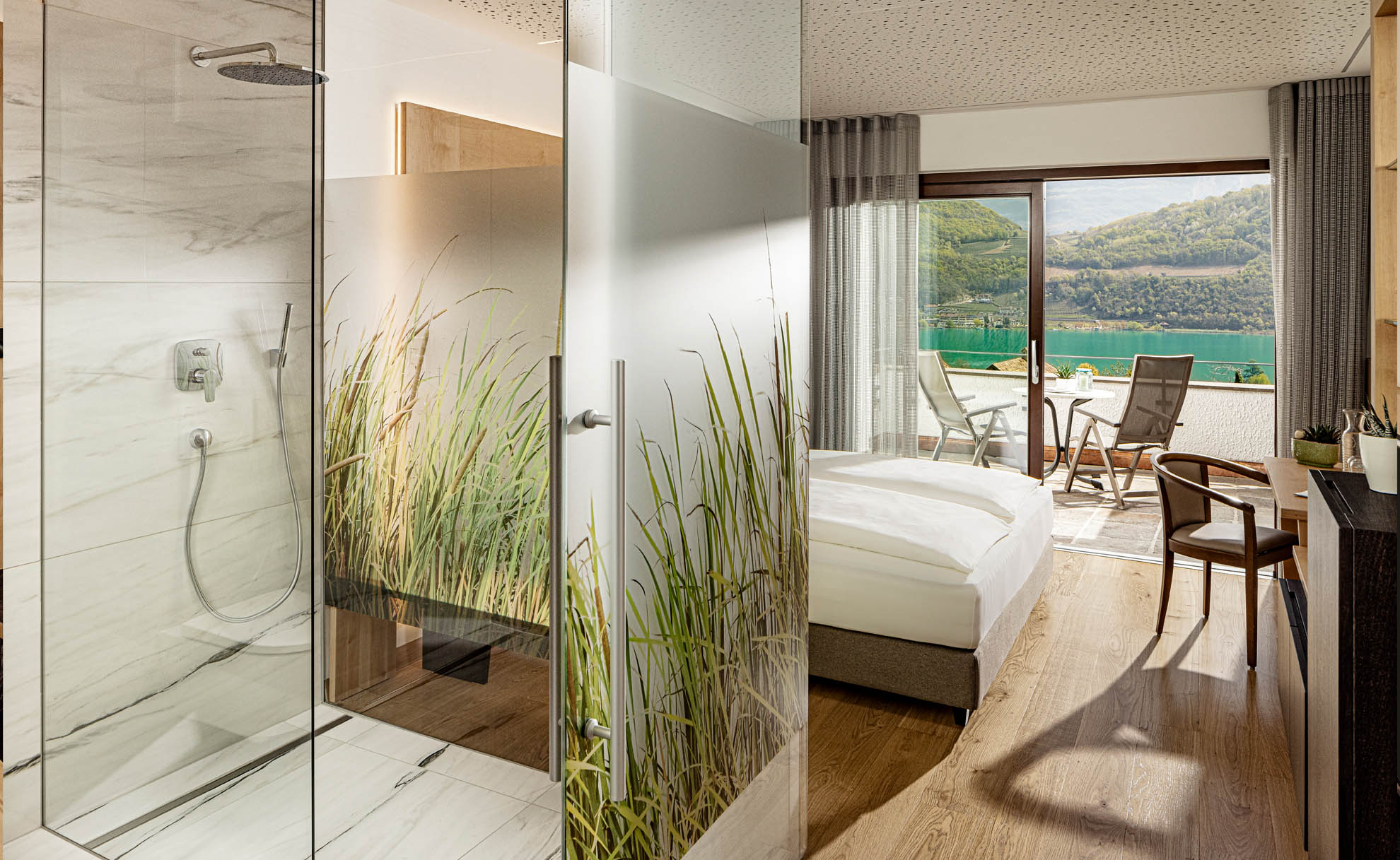 Modernes Hotelzimmer mit Doppelbett, gl&auml;serner Duschwand mit Gr&auml;sermotiv und bodentiefer Fensterfront mit Blick auf den See u