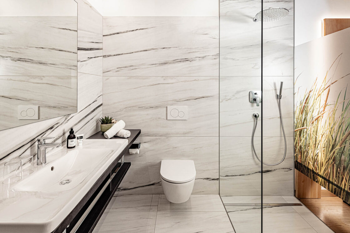 Marmorbad im Hotel mit bodengleicher Glasdusche, wandhängendem WC, offener Ablage mit Waschbecken und Handtüchern.