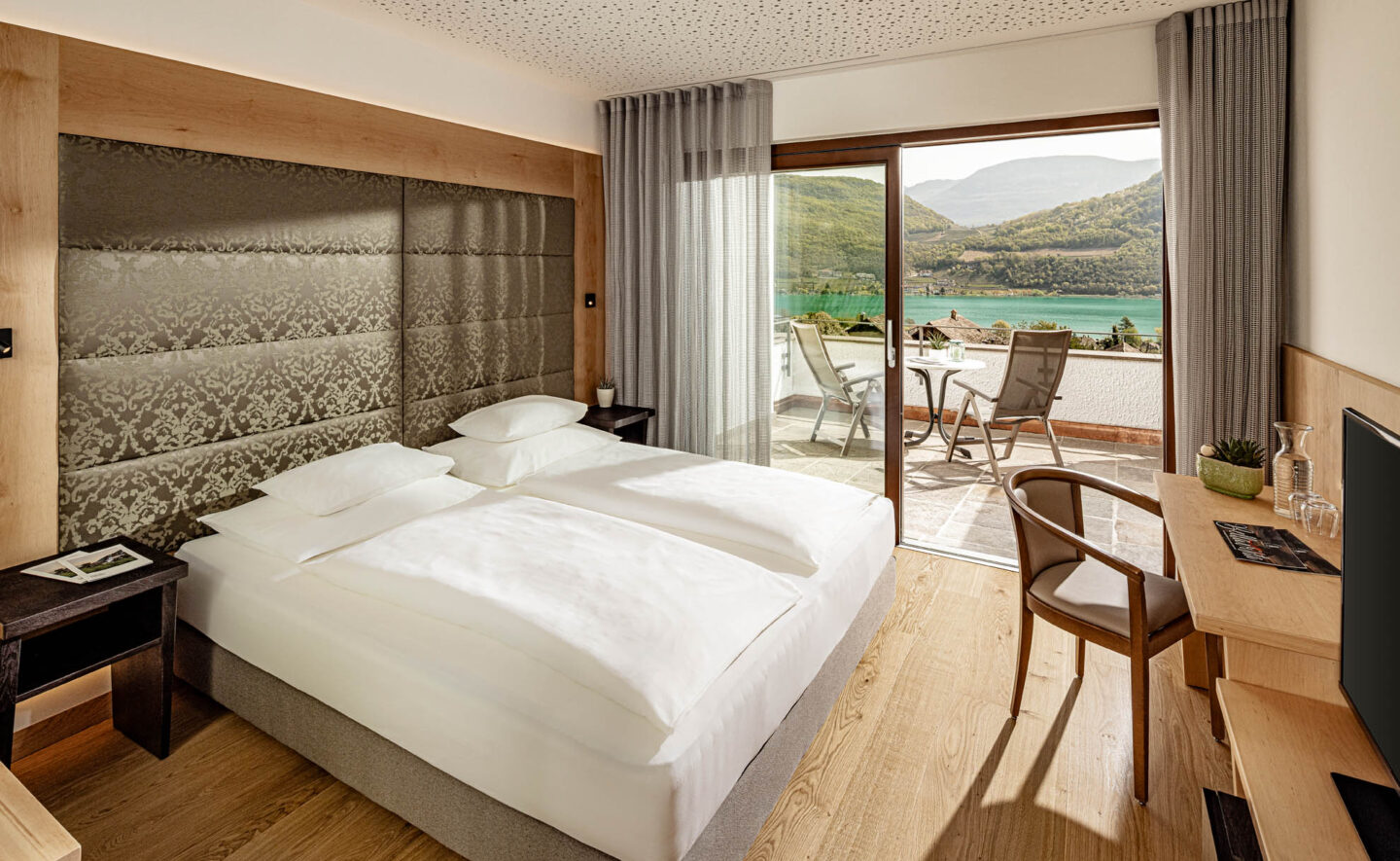 Doppelzimmer mit Bett, gepolstertem Kopfteil, Nachttischlampen und Balkon mit St&uuml;hlen und Blick auf den Kalterer See