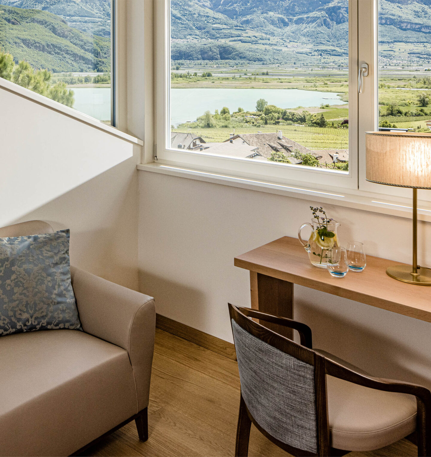 Hotelzimmer-Ecke mit Sessel, Schreibtisch, Lampe und Stuhl; gro&szlig;es Fenster mit Blick auf See und H&uuml;gel.