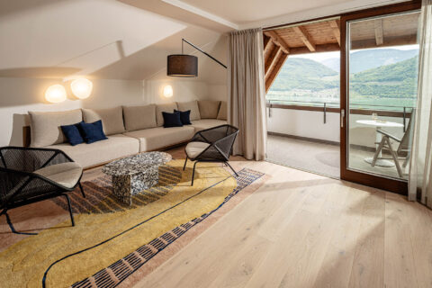 Wohnzimmer mit Sofa, Couchtisch, Stehlampe und Holzbalkendecke; geöffnete Balkontür mit Blick auf Hang und Kalterer See.