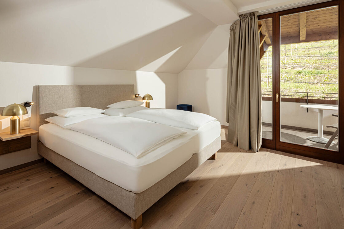 Doppelbett in heller Suite mit schräger Decke, Nachttischen, Holzfußboden und geöffneter Balkontür mit Vorhang.