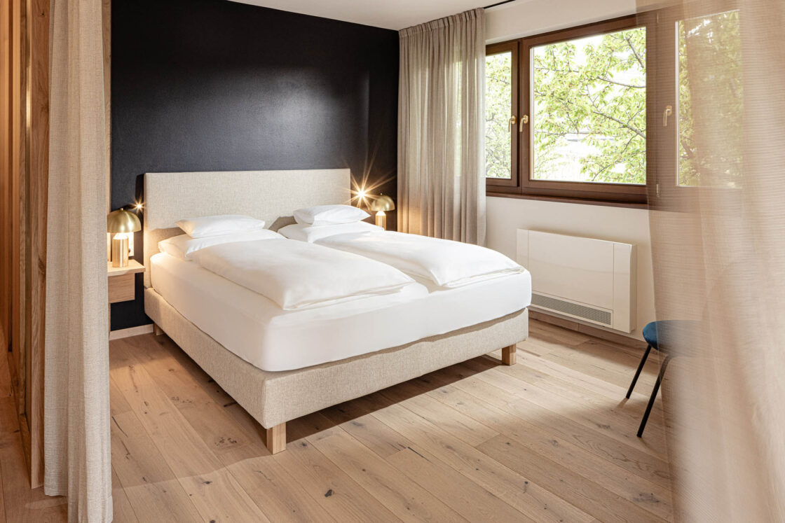 Hotelzimmer (See Suite) mit Doppelbett (zwei Decken), Nachttischlampe, Holzboden und Fenster mit Blick auf Bäume