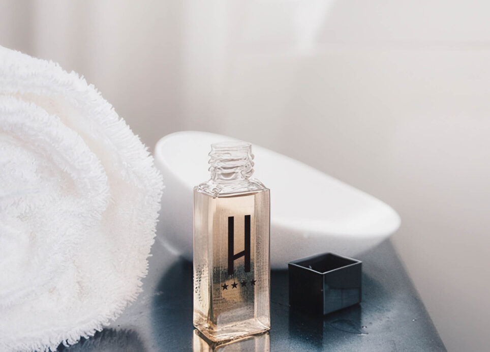 Glasflasche mit klarem Inhalt und H‑Logo auf dunkler Ablage; schwarzer Deckel daneben, weiße Handtuchkante und unscharfe Seif