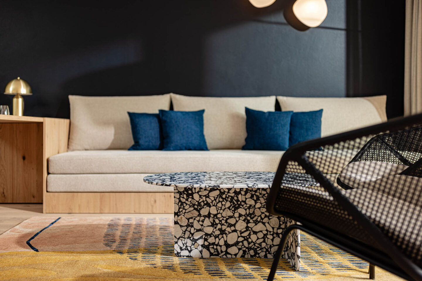 Beiges Sofa mit drei blauen Kissen, schwarzer gemusterter Couchtisch, Rattanstuhllehne, Teppich und Wandlampe.