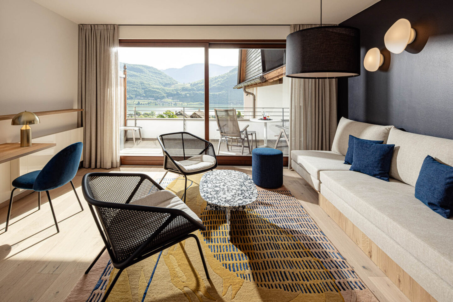 Hotelzimmer-Lounge mit zwei Stühlen, Sofa, Couchtisch und großem Panoramafenster mit Blick auf See und Berge.