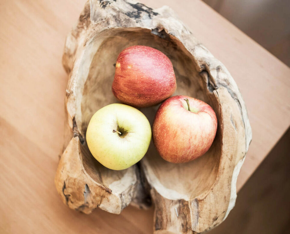 Ausgehöhlte Holzschale mit drei Äpfeln (zwei rot, einer grünlich-gelb) auf heller Oberfläche