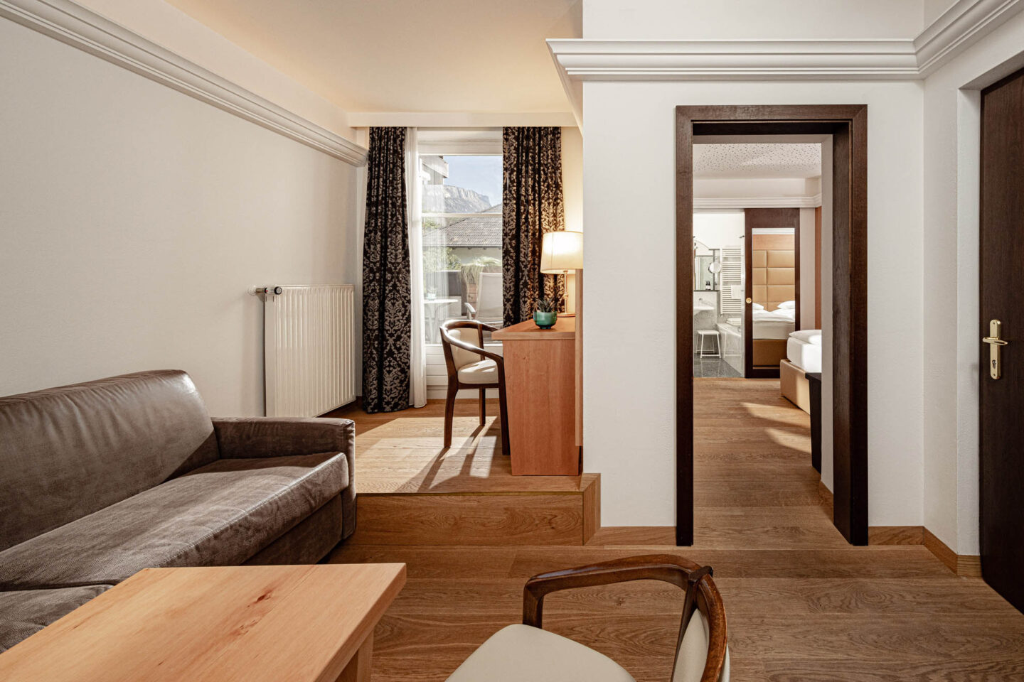 Suite mit Wohnbereich und angrenzendem Schlafzimmer: Sofa, Holztisch mit Stühlen, Stehlampe, Vorhänge und sichtbar gemachtes