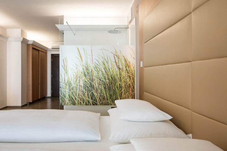 Doppelzimmer mit weißer Bettwäsche, mehreren Kissen, gepolstertem Kopfteil und Wandbild mit hoch wachsendem Gras.