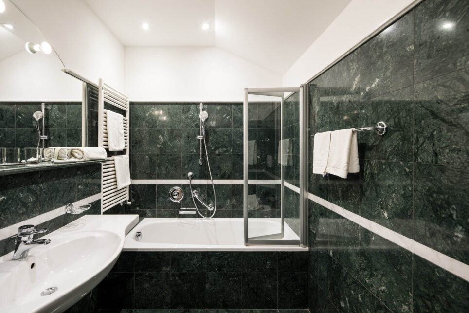 Badezimmer mit Badewanne und Glas-Duschabtrennung, dunkle grüne Marmorwände, Wandhandtuchhalter und Rohrheizkörper.