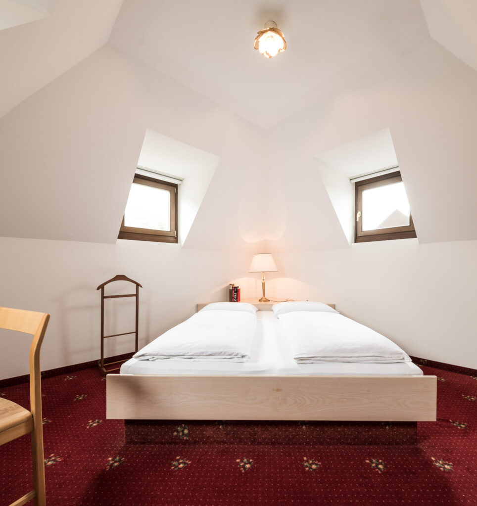 Schlafzimmer in Mansarde mit Doppelbett, zwei Giebelfenstern, Nachttisch mit Lampe, Stuhl, Gepäckablage und rotem Teppich