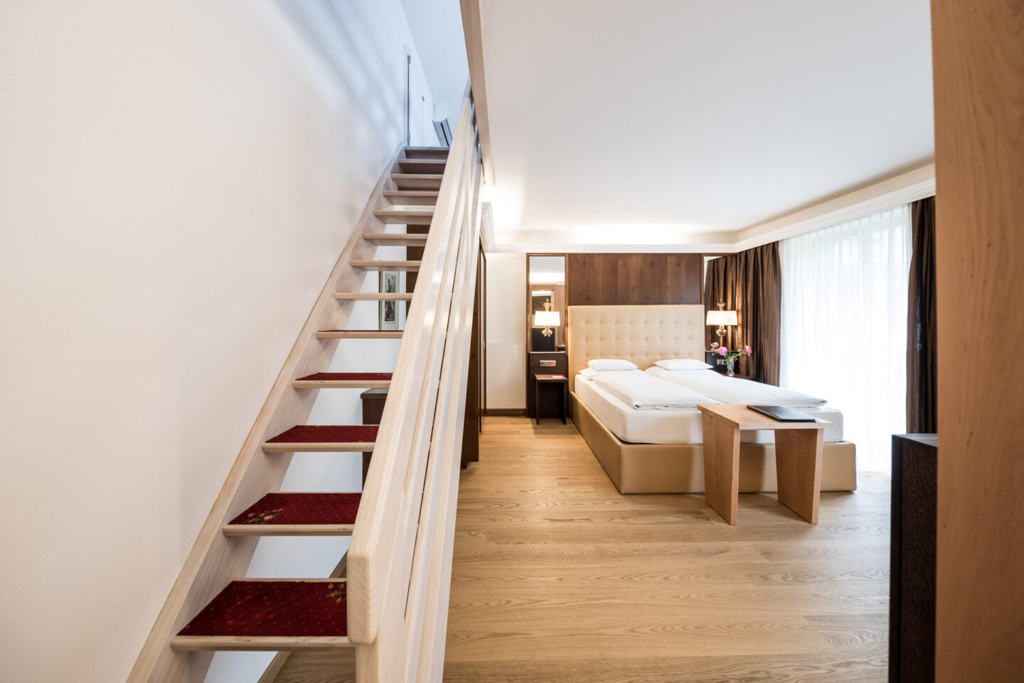 Suitezimmer mit offener Holztreppe links, Doppelbett, Holzfußboden, Sitzbank und großen Fenstern mit Vorhängen