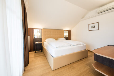 Doppelbett in heller Suite mit schräger Decke, zwei Nachttischen mit Lampen, Parkettboden und geschlossenem Vorhang