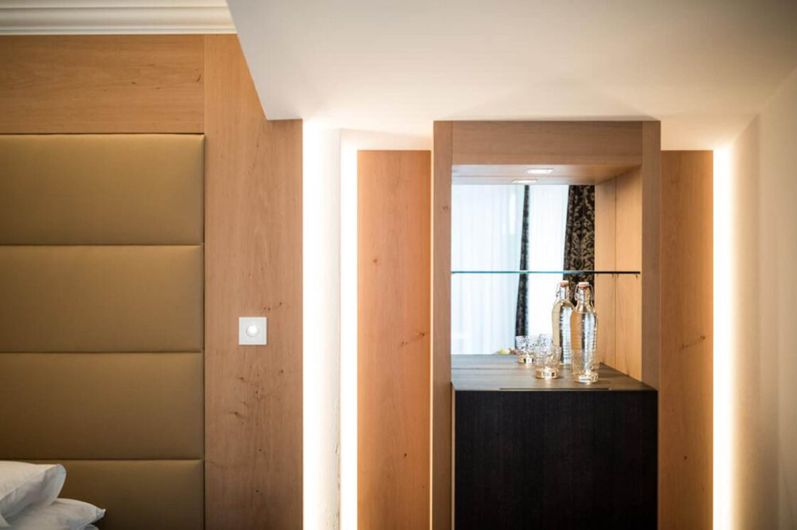 Holzgetäfelter Eingang eines Hotelzimmers, Lichtschalter, Durchgang zu hellem Badfenster, schwarzer Schrank mit Glasvasen