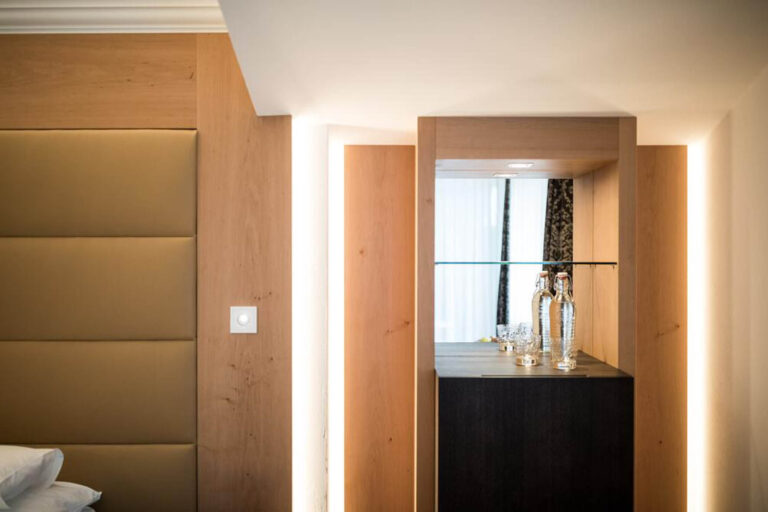 Holzgetäfelter Eingang eines Hotelzimmers, Lichtschalter, Durchgang zu hellem Badfenster, schwarzer Schrank mit Glasvasen