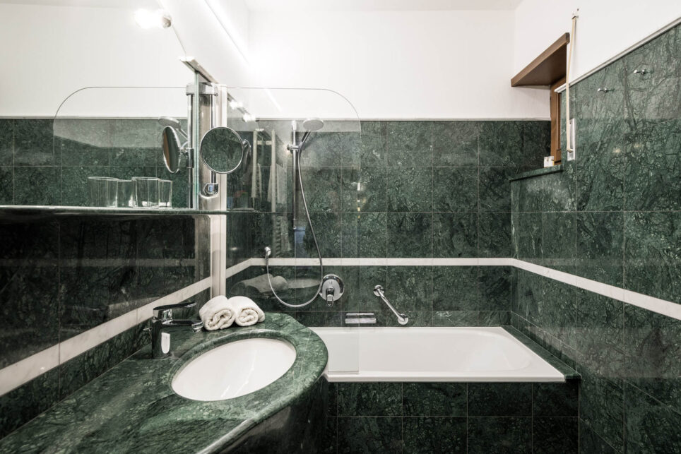 Hotelbadezimmer mit dunkelgrüner Marmorverkleidung, Waschbecken, Spiegel, verstellbarem Schminkspiegel, Badewanne mit Dusche.