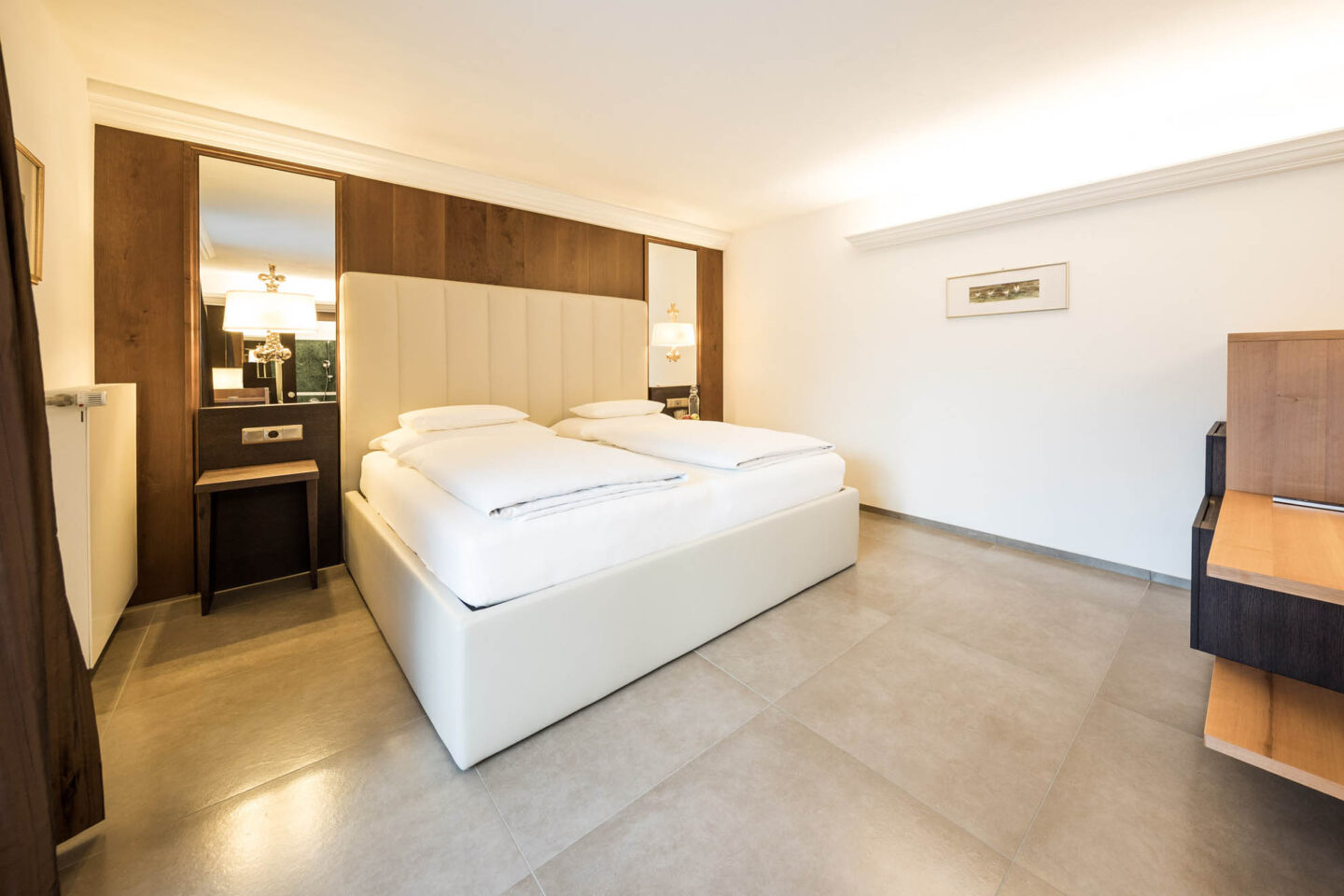 Zimmer mit zwei zusammenstehenden Matratzen, gepolstertem Kopfteil, Nachttischlampe, hellem Fliesenboden und kleinem Wandbild