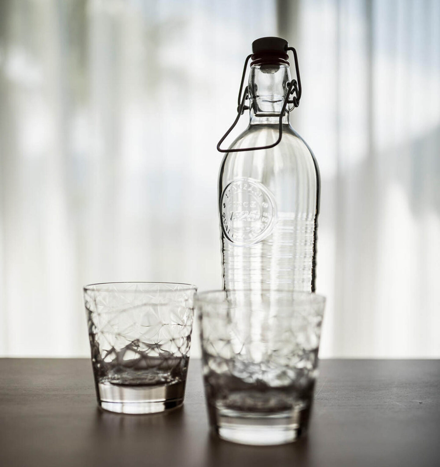 Karaffe mit Bügelverschluss neben Trinkglas auf dunklem Tisch vor durchscheinenden Vorhängen.