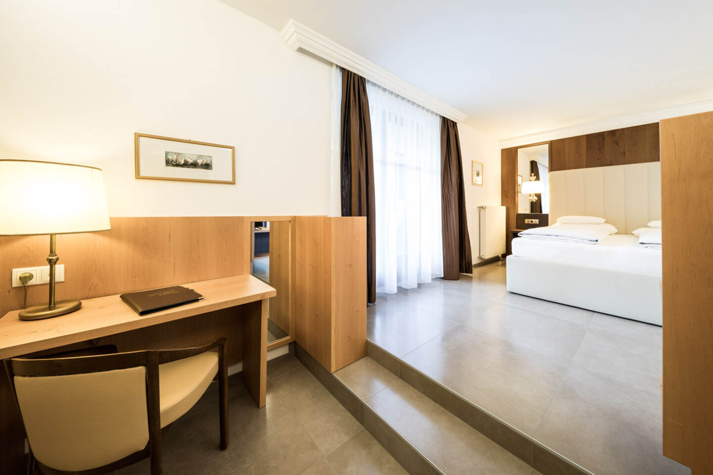 Helles Hotelzimmer Weinberg mit Schreibtisch und Stuhl links, bodentiefer Gardine, erhöhtem Doppelbett und Holzdetails.