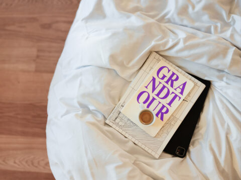 Wei&szlig;e, zerknitterte Bettw&auml;sche auf Holzfu&szlig;boden; darauf ein geschlossenes Buch mit buntem Schriftzug und ein dunkles Rechteck