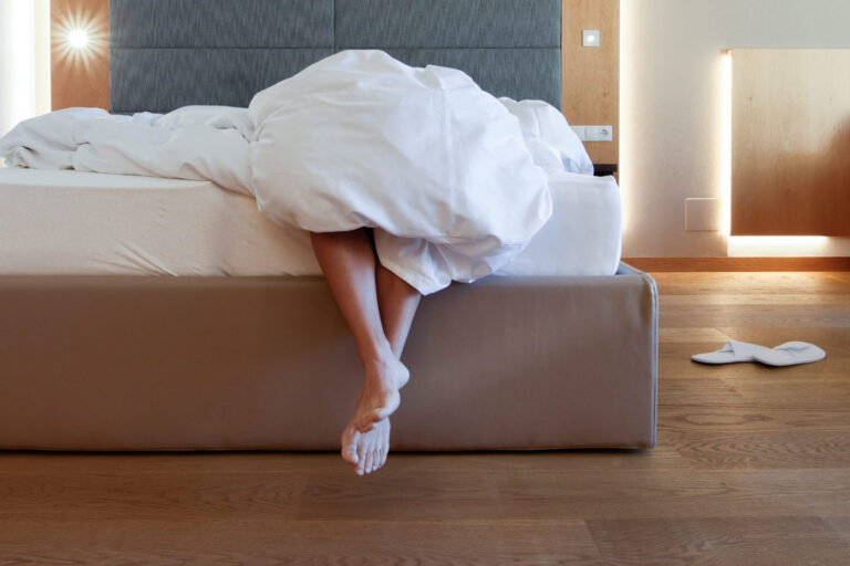 Beine einer Person ragen unter zerwühlter weißer Bettdecke über die Bettkante in einem Hotelzimmer mit Holzboden.