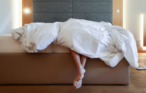 Zerwühltes Hotelbett mit weißer Decke; zwei nackte Füße ragen unter der Decke hervor, Stoff-Kopfteil und Wand im Hintergrund.