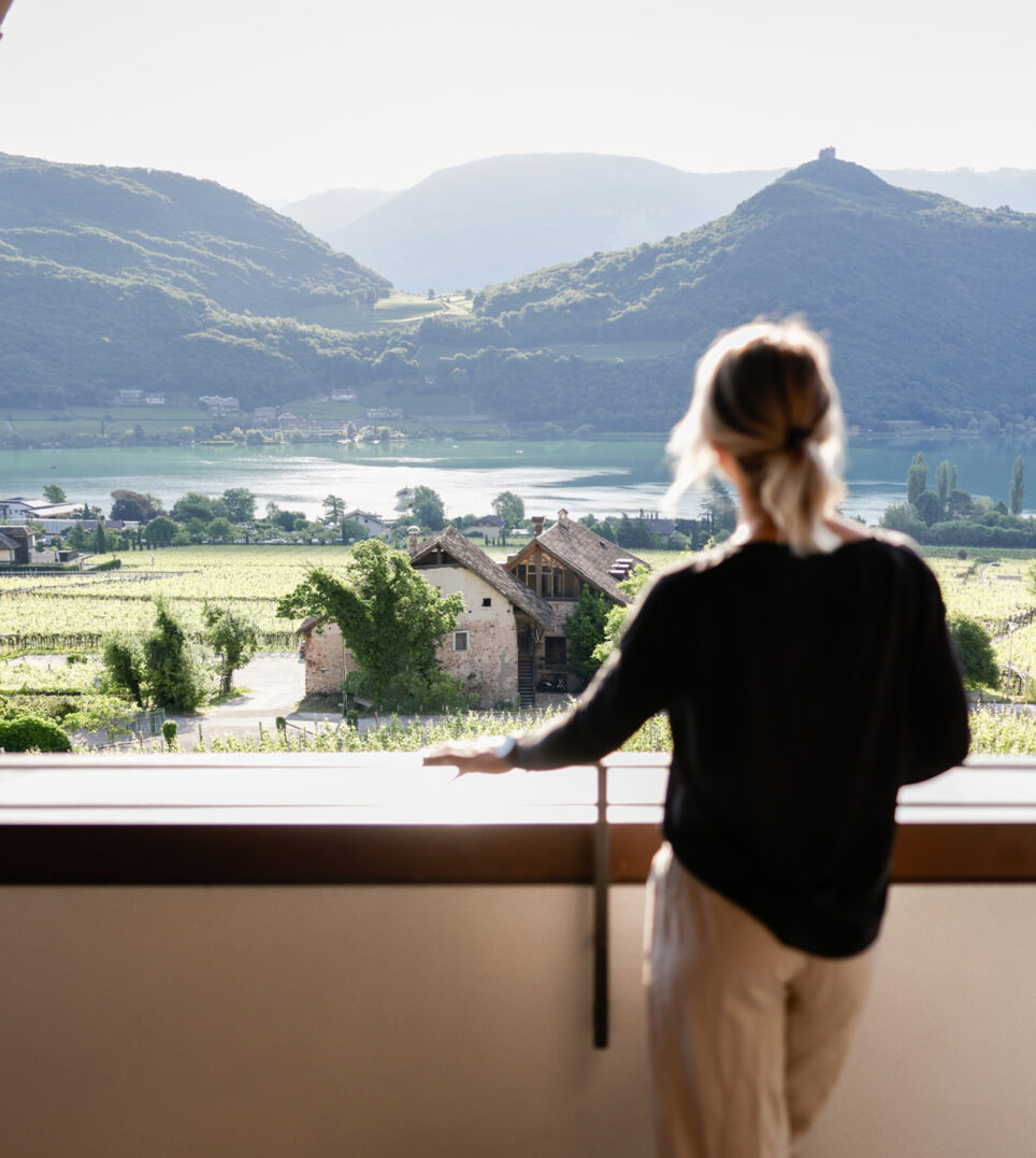 Person mit zusammengebundenem Haar steht an einem Balkon und blickt auf See, Felder und sanfte Hügel