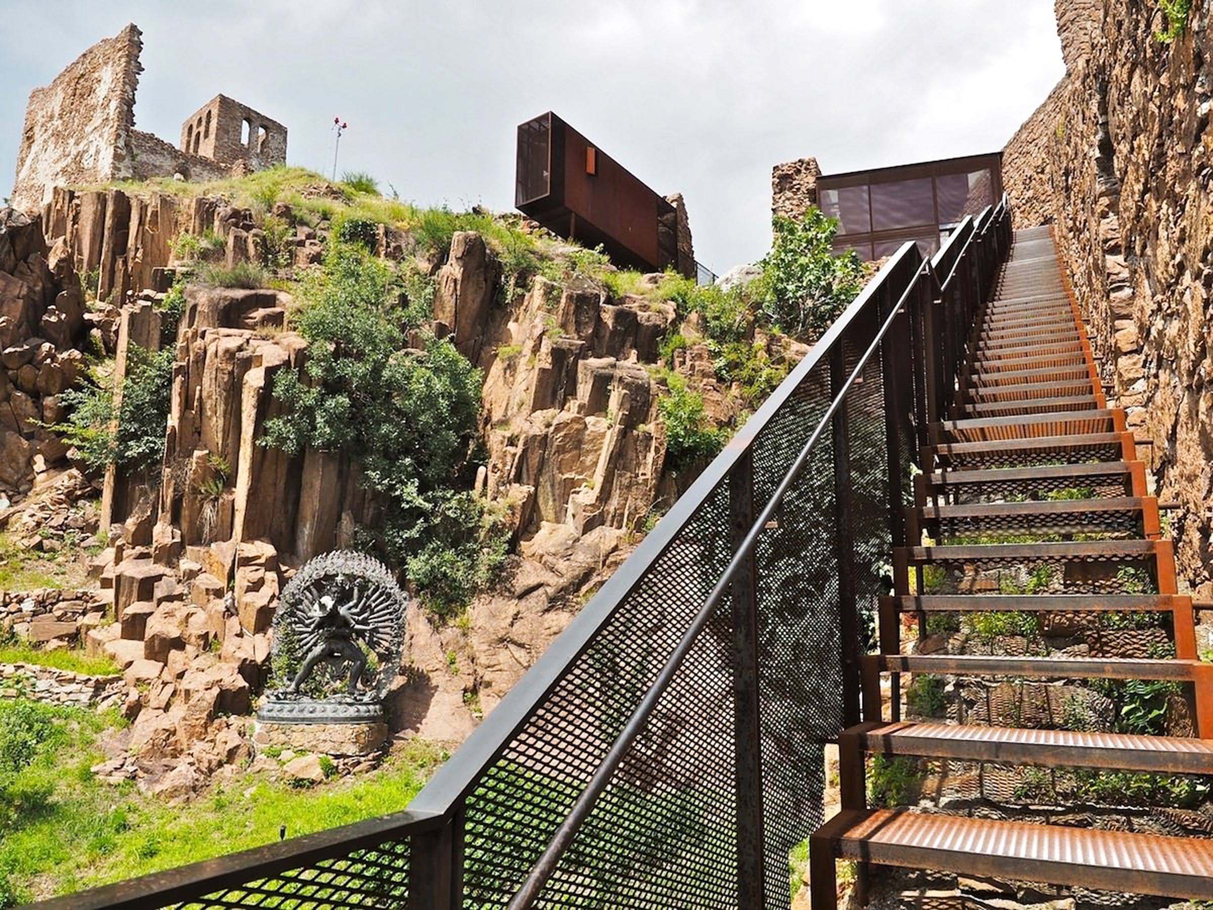 Metalltreppe an steiler Felswand mit Geländer; darunter Skulptur und bewachsene Mauern, Gebäude auf dem Felsplateau.