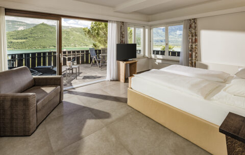 Helle Suite mit großem Bett, Sofa und TV, bodentiefe Fenster zu Balkon mit Tisch und Stühlen; Blick auf Bäume.