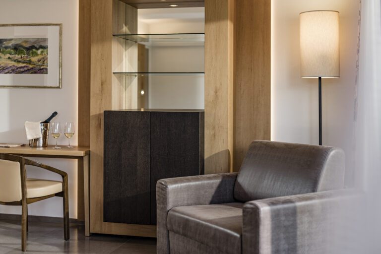 Ecke einer Suite mit grauem Polstersessel, schwarzem Schrank mit Glasfächern, Holzverkleidung, kleinem Tisch und Wandlampe