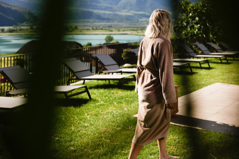 Rückenansicht einer Person im Bademantel auf Rasen, geht zu Liegestühlen am Ufer; davor dunkle Pflanzen als Rahmen.