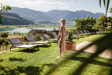 Frau in lockerem Kleid steht auf Hotelwiese neben Liegest&uuml;hlen und kleinem Koffer, Blick zum See und den Bergen.