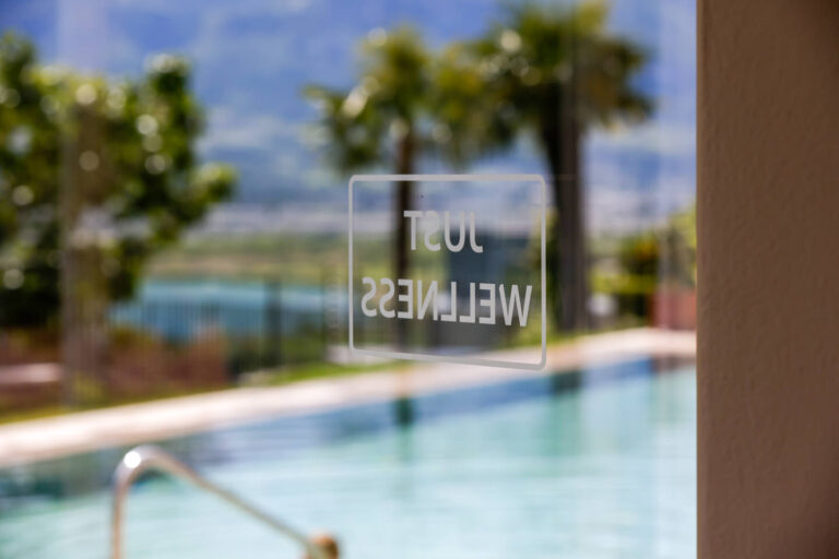 Glastür mit spiegelnder Beschriftung HOTEL WELLNESS, im Hintergrund Außenpool, Palmen und der Kalterer See vor Bergen